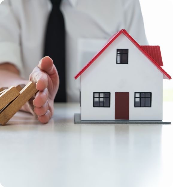Person in a shirt and tie stopping falling wooden blocks next to a small model house with a red roof.