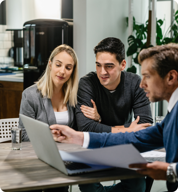 A man and woman attentively listening to a professional explaining details on a laptop at a table in a modern office.