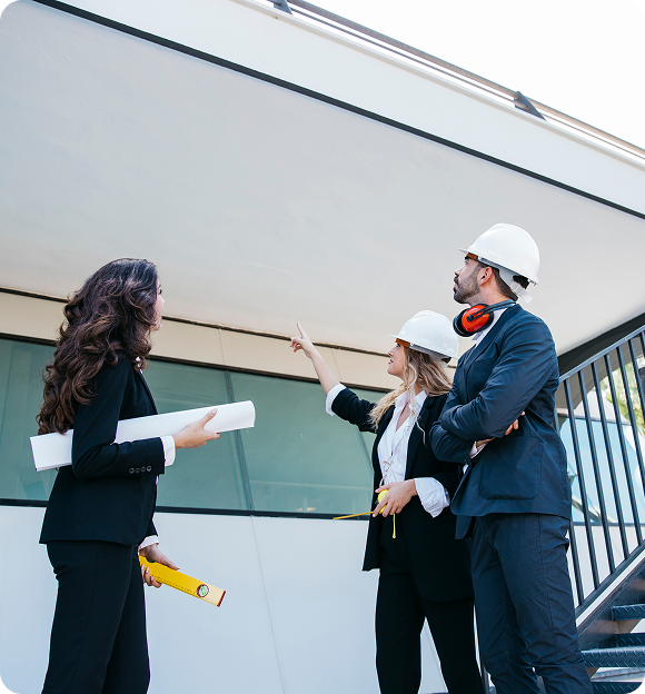 Three architects or engineers wearing hard hats and business attire inspecting a building, one woman pointing upwards while holding a measuring tape and another woman holding rolled blueprints.
