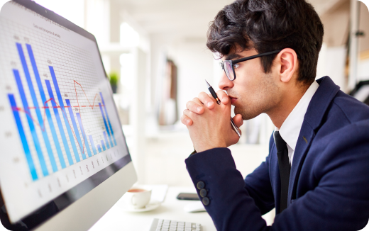 Man in a suit and glasses intently analyzing a bar and line chart on a desktop computer screen.