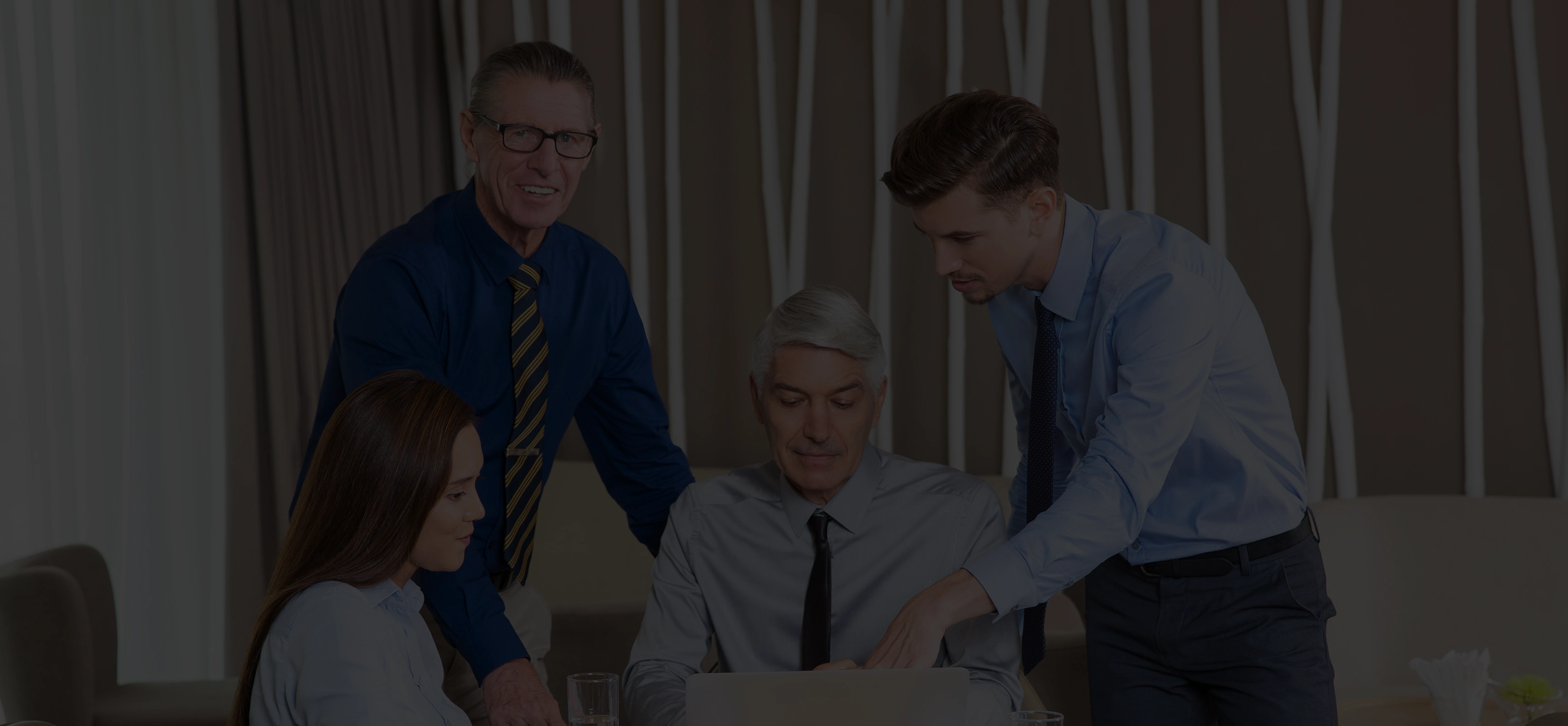 Four business professionals, two men and two women, gathered around a laptop discussing work in an office.