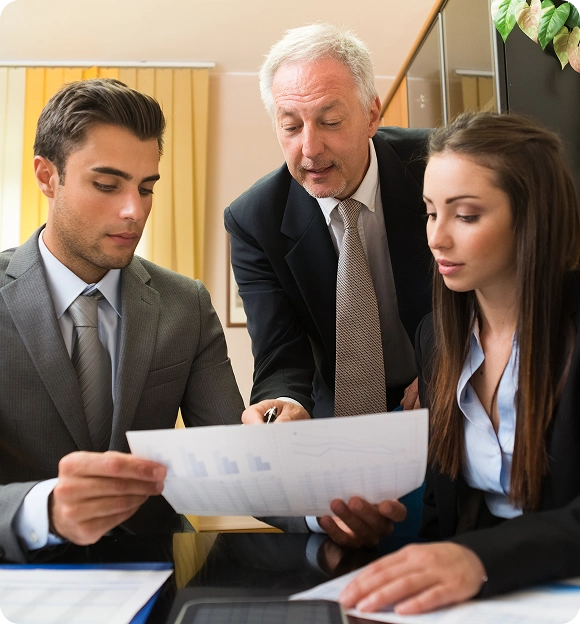 Three business professionals in suits reviewing a document with charts in an office setting.