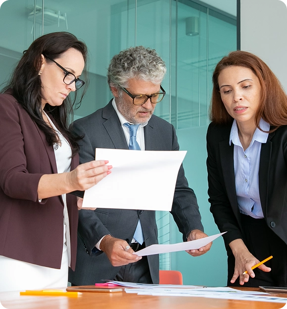 Three business professionals standing around a table reviewing documents in an office with glass walls.