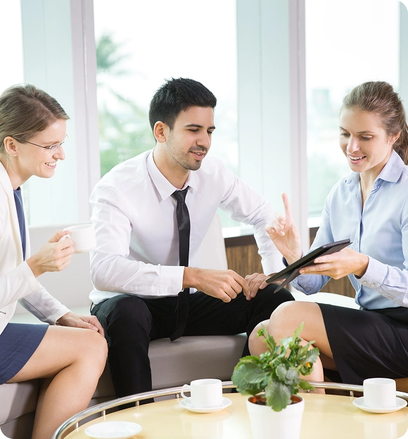 Three business professionals sitting on a couch, with one woman showing information on a tablet to the others in a bright office setting.
