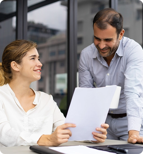 Two colleagues smiling and discussing over a document in an office setting.