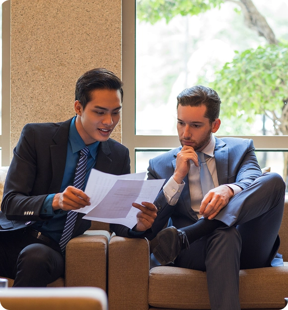 Two businessmen in suits sitting on a couch reviewing documents together near a window.
