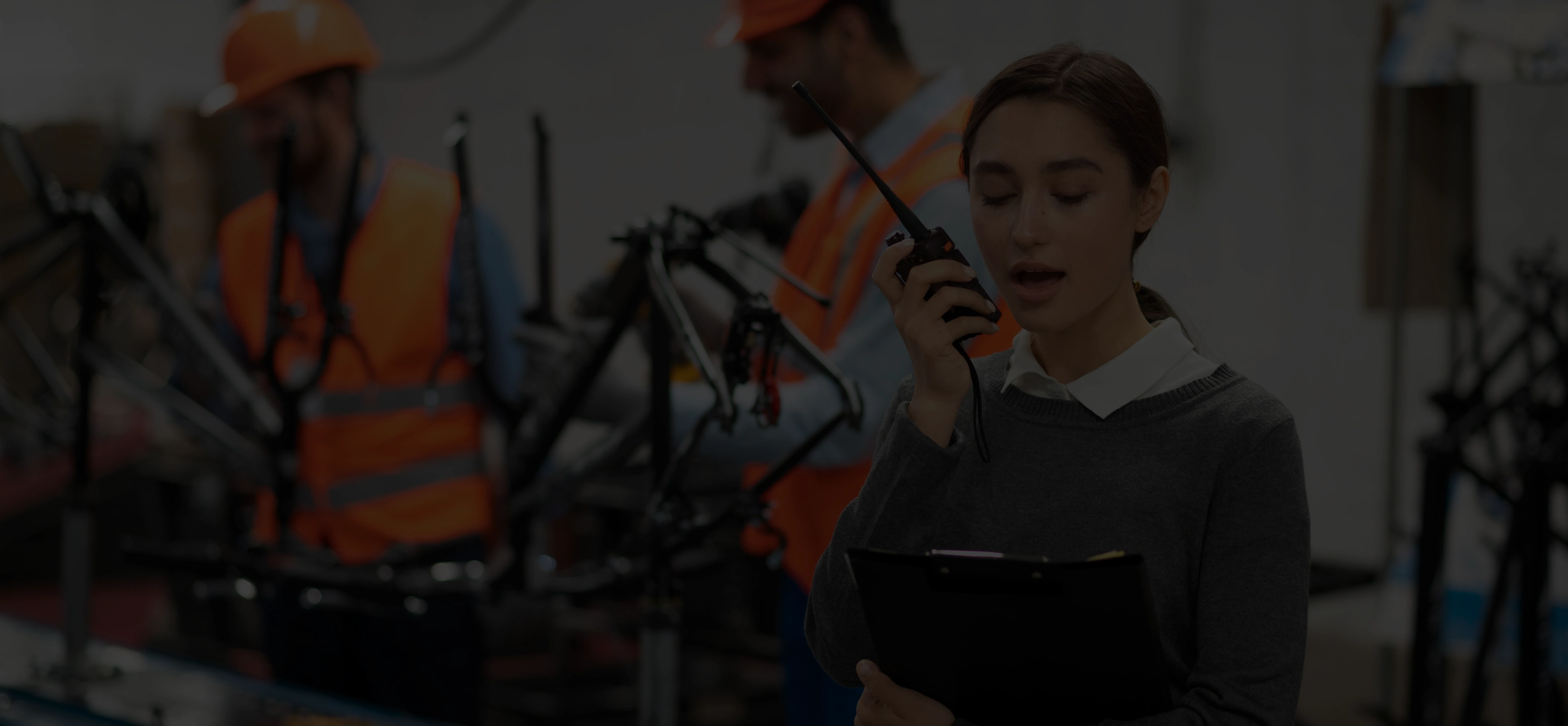 Woman holding a clipboard and speaking into a walkie-talkie in a factory setting with workers assembling bicycles in the background.