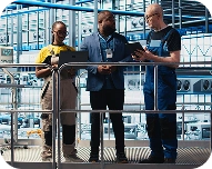 Three industrial workers wearing safety gear discussing a tablet in a factory setting with metal frameworks in the background.
