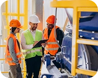 Three construction workers wearing hard hats and safety vests discussing documents in an industrial setting.
