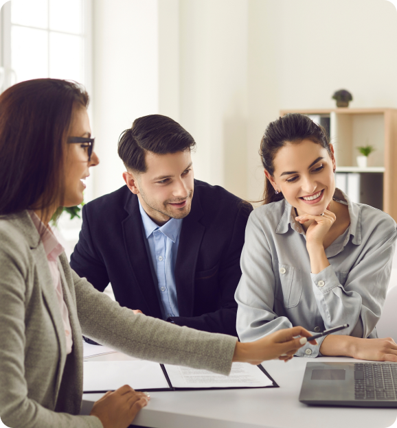 Three professionals in office attire engaged in a discussion around a laptop, with two smiling and one pointing at the screen.
