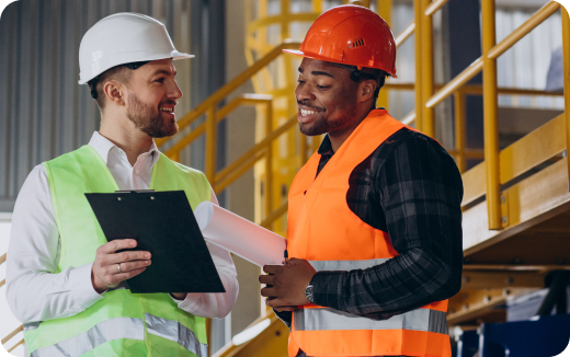 Two construction workers in safety vests and helmets discussing a clipboard and blueprint inside an industrial building.