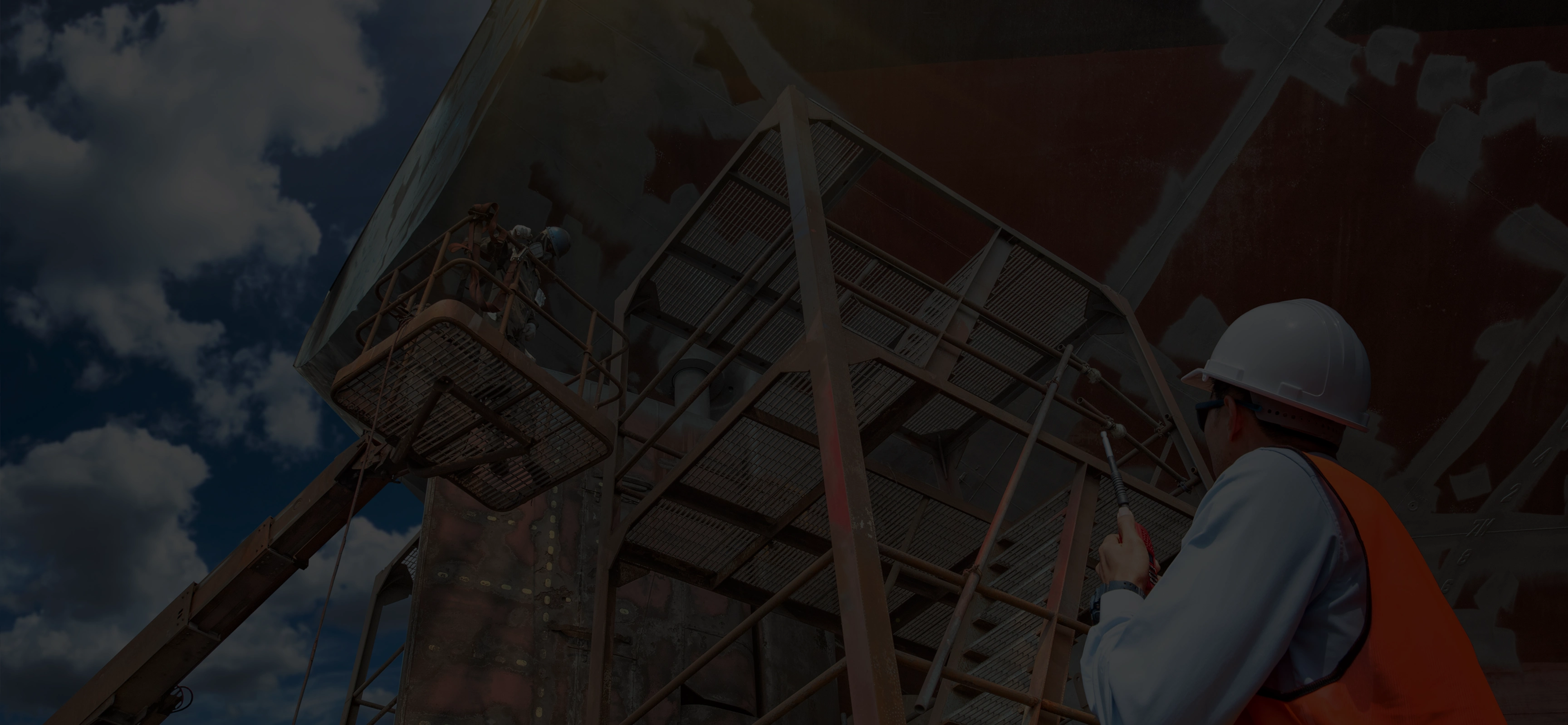Construction worker in white hard hat and orange vest inspecting or working on elevated industrial metal structure using a tool.