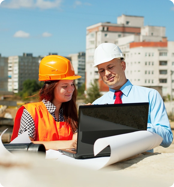 Two construction professionals wearing helmets and safety gear reviewing plans on a laptop at a construction site with buildings in the background.
