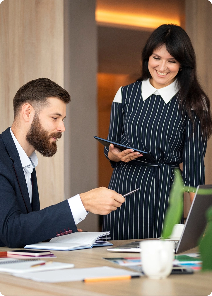 Man in suit seated at desk pointing at a laptop screen with a pen while a woman in a striped dress holds a tablet and smiles standing beside him.