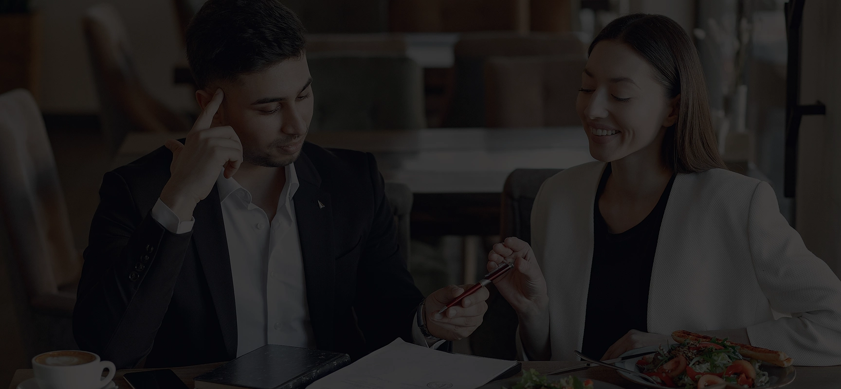 Two business professionals sitting at a restaurant table discussing documents with a pen and a salad plate nearby.