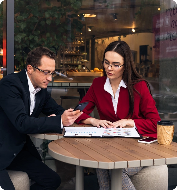 Two professionals in business attire reviewing documents and using a smartphone at a round wooden table outside a café.