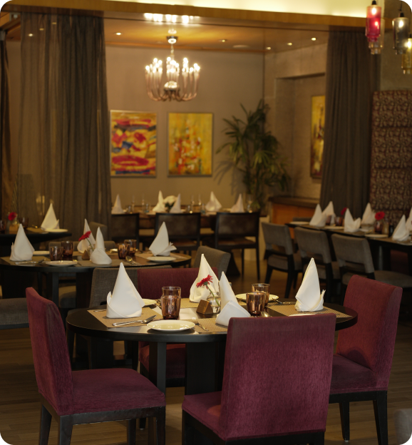Cozy restaurant dining area with round tables set with white napkins, brown glasses, and pink chairs under warm lighting and abstract wall paintings.