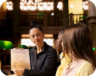 A professional woman holding and explaining a clipboard to two attentive women in a warmly lit office.