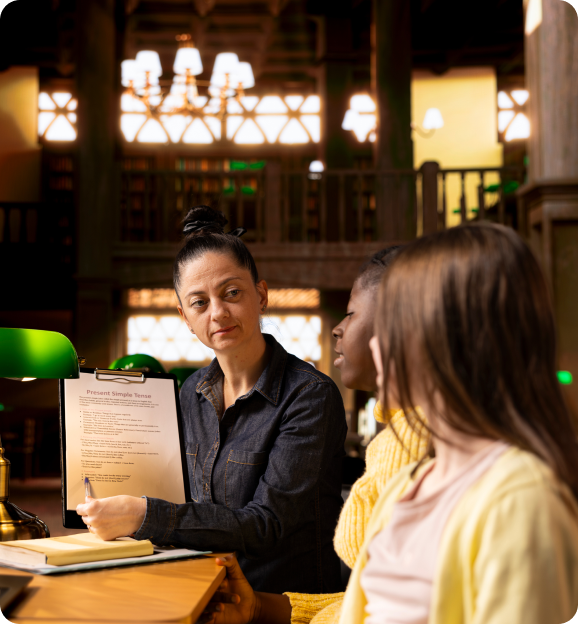A tutor explaining a document titled 'Present Simple Tense' to two students in a warmly lit library setting.