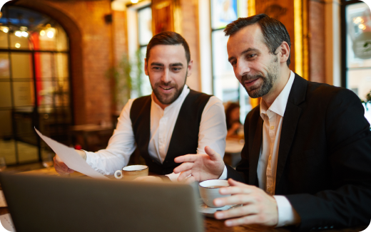 Two men in business attire discussing documents over coffee while looking at a laptop in a cozy cafe.