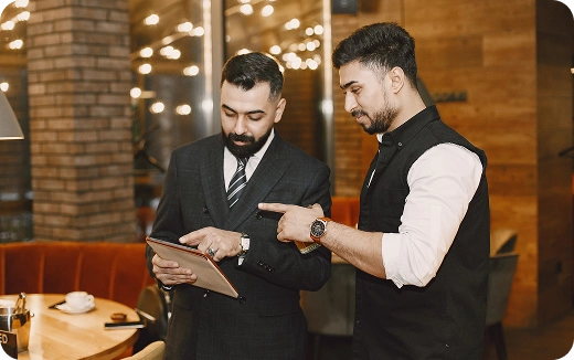 Two men in business attire reviewing content on a tablet inside a warmly lit cafe.