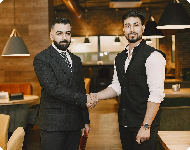 Two men shaking hands in a warmly lit modern restaurant, one wearing a suit and tie, the other in a black vest and white shirt.