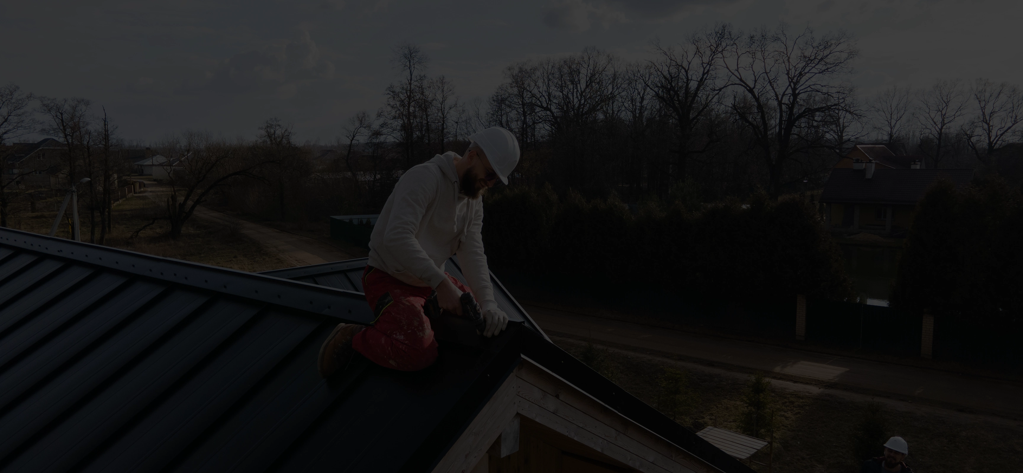 Construction worker wearing a white hard hat and gloves installing a black metal roof panel on a house.