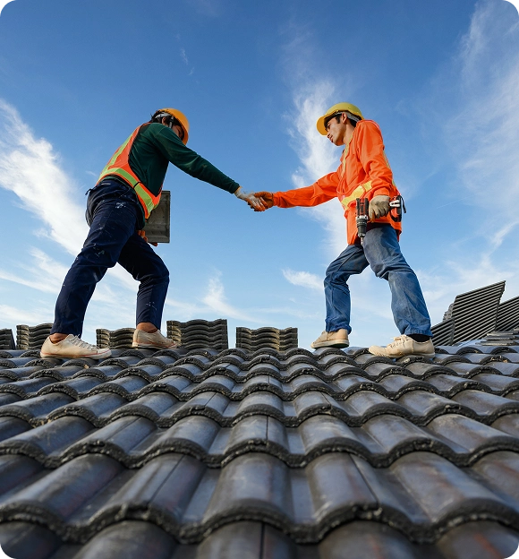 Two construction workers wearing safety gear shaking hands while standing on a tiled roof under a partly cloudy sky.