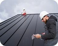 Two workers wearing white helmets inspecting a large sloped metal roof under cloudy sky.