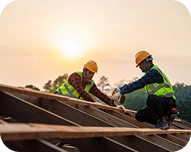 Two construction workers wearing helmets working on a wooden roof frame at sunset.
