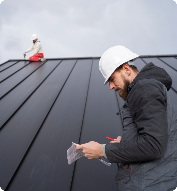 Construction worker in white hard hat and gray jacket examining papers on a black metal roof with another worker in the background.