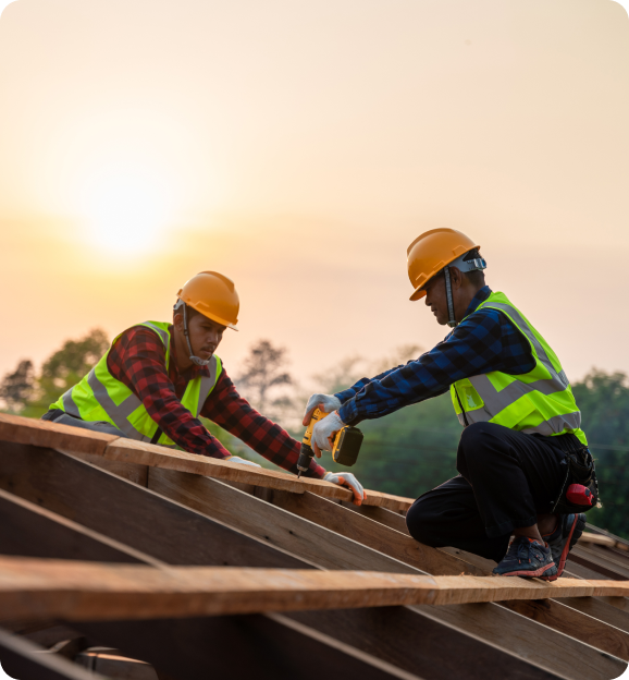 Two construction workers in safety vests and helmets working on wooden roof framing during sunset.