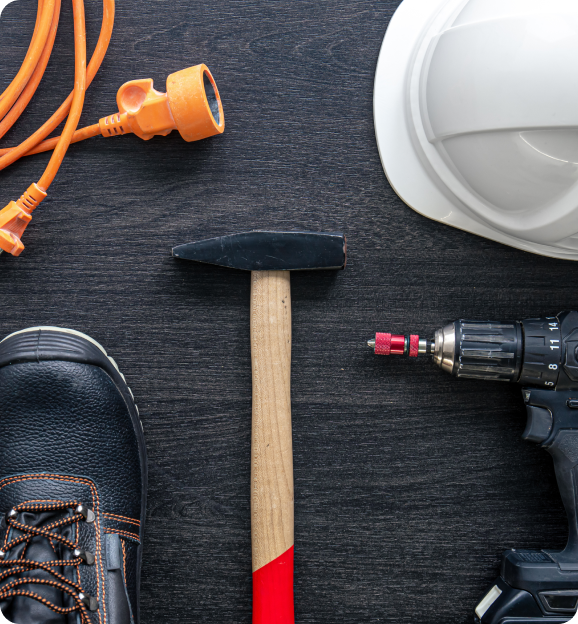 Work boots, orange extension cord, hammer with wooden handle, white safety helmet, and a cordless drill on a dark wooden surface.