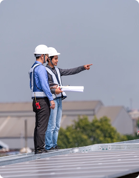 Two construction workers in safety helmets and vests reviewing plans and pointing while standing on a rooftop.