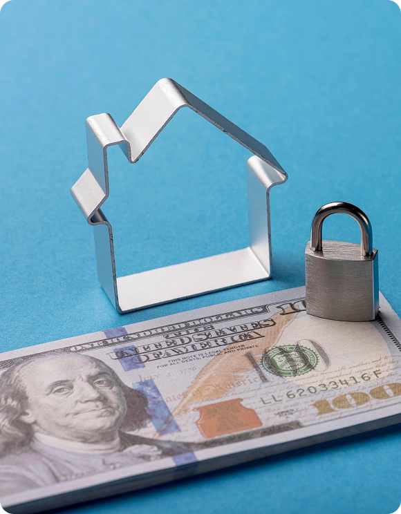 Metal house-shaped cookie cutter and small silver padlock placed on a stack of US 100 dollar bills on a blue background.