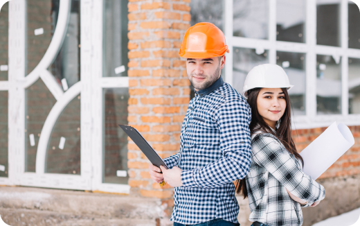 Man and woman wearing hard hats and checkered shirts standing back-to-back outside a building with rolled-up blueprints and clipboard.