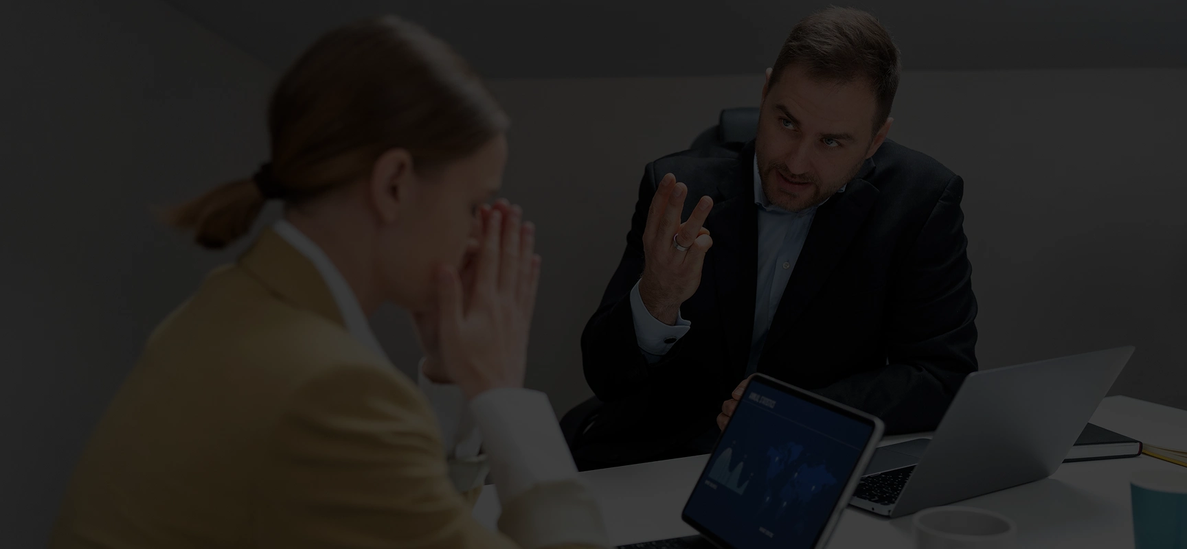 Two business professionals having a serious discussion at a desk with laptops and documents.