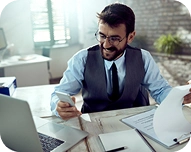 Man in glasses and vest smiling while reviewing documents and holding cash at a desk with a laptop.