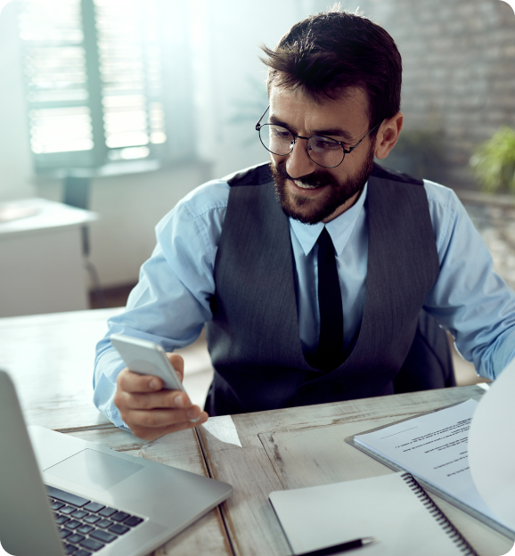 Smiling man in glasses and vest sitting at desk, looking at smartphone with laptop and documents nearby.