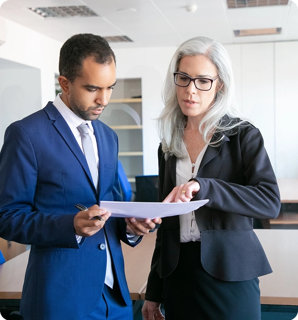 A businesswoman with gray hair and glasses discussing documents with a man in a blue suit in an office.