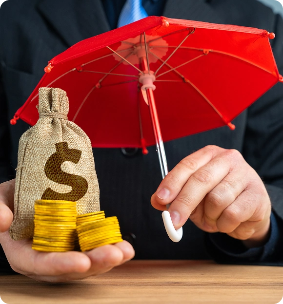 Person in suit holding a small red umbrella over a burlap money bag with a dollar sign and stacks of gold coins in their palm.