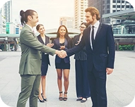 Two business professionals shaking hands outdoors with three colleagues standing behind them in a city setting.
