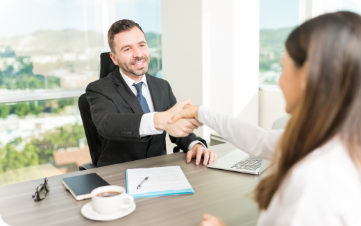 Man in suit shaking hands with woman across a desk in a bright office with a city view.