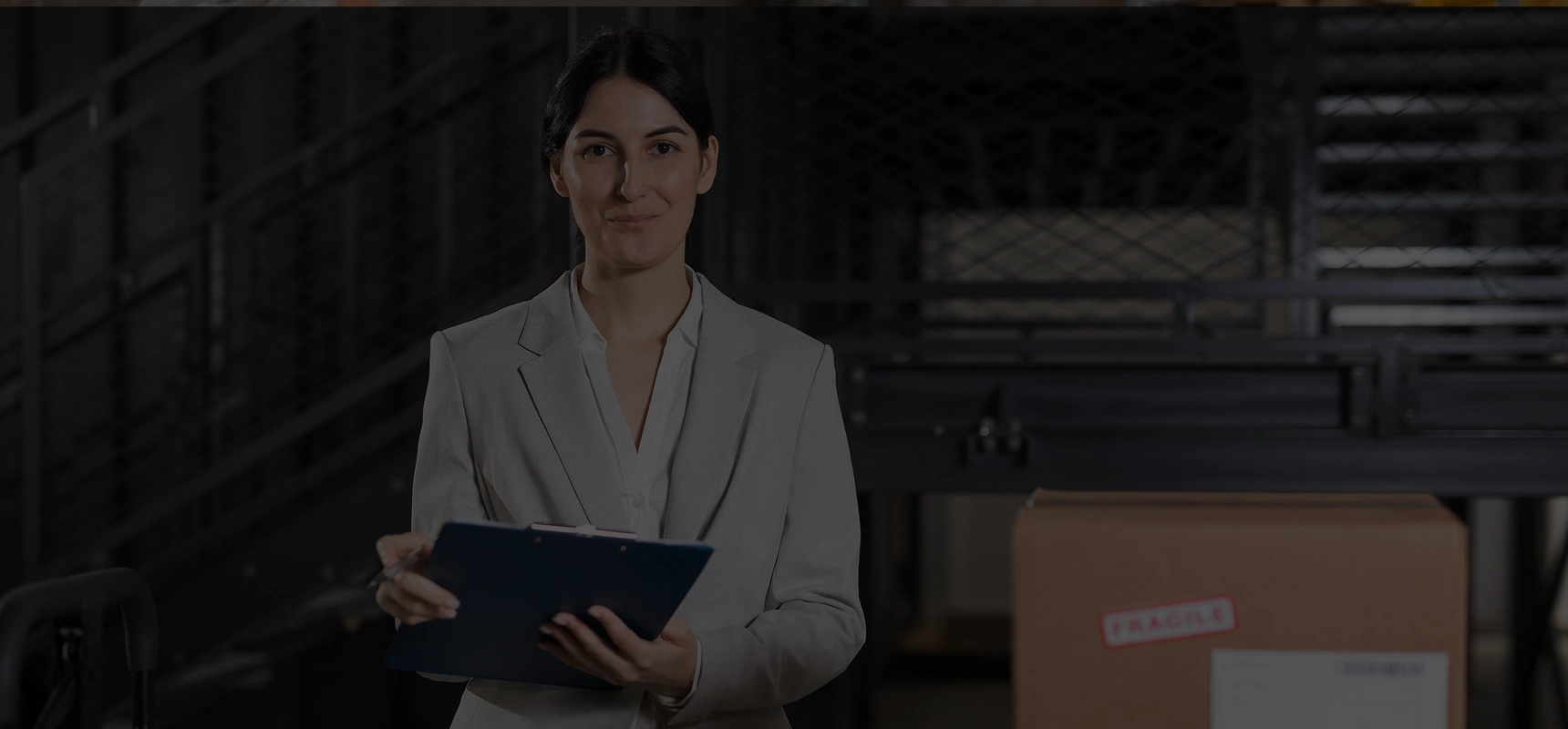Professional woman in a light gray blazer holding a clipboard in a warehouse with a cardboard box labeled fragile.