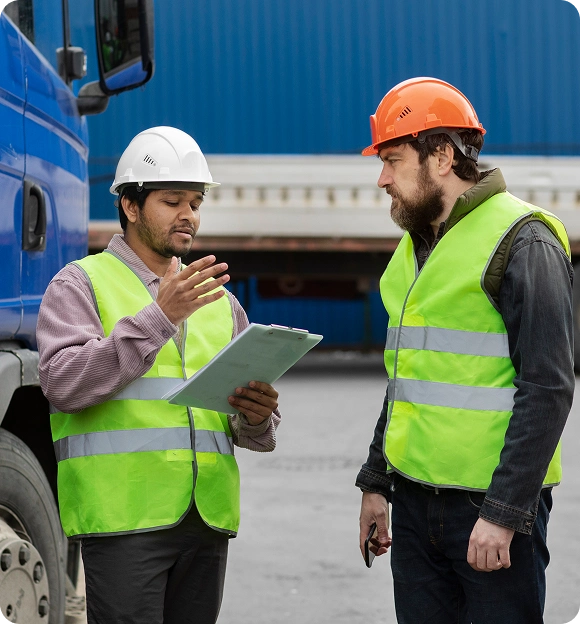 Two construction workers in safety vests and helmets discussing with a clipboard near a blue truck.