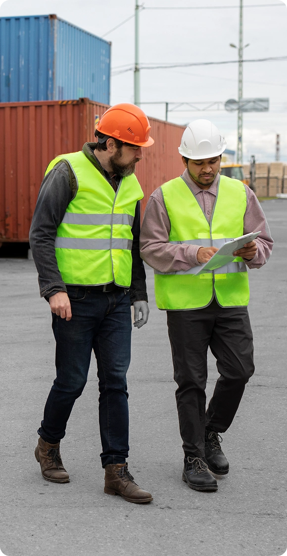 Two men wearing high-visibility vests and safety helmets reviewing documents at a shipping container yard.