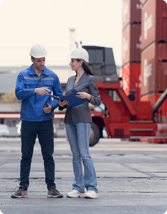 Man and woman wearing white safety helmets reviewing documents in an industrial shipping container yard.