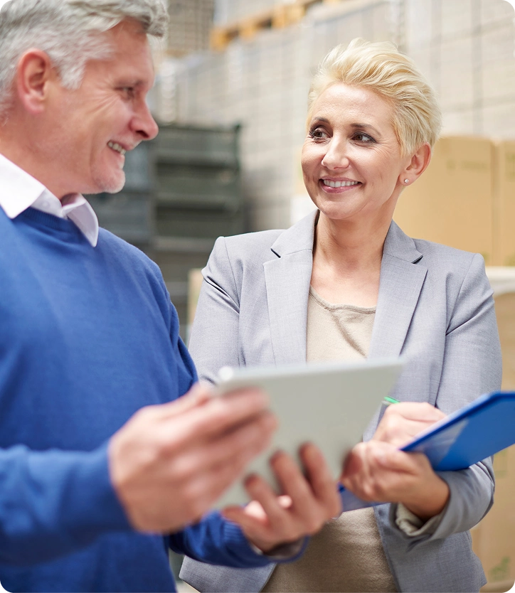Two colleagues smiling and discussing while holding a tablet and clipboard in a warehouse setting.