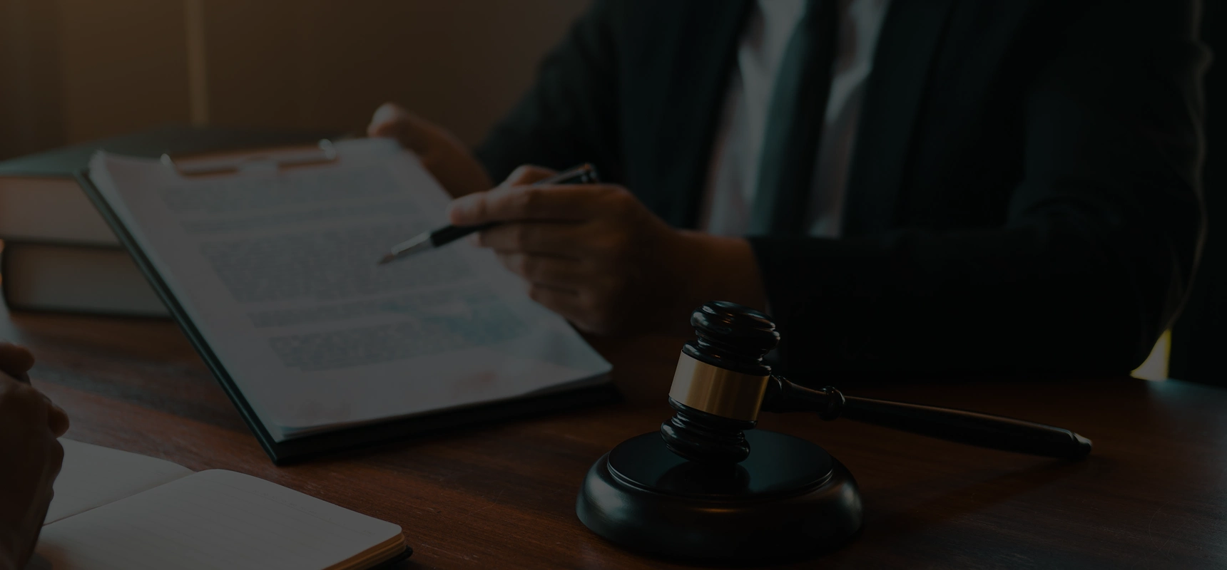 Person in suit holding a pen and pointing to a document on a clipboard beside a judge's gavel on a wooden table.