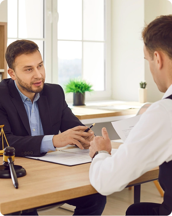 Two men in a professional office setting discussing documents across a wooden desk.
